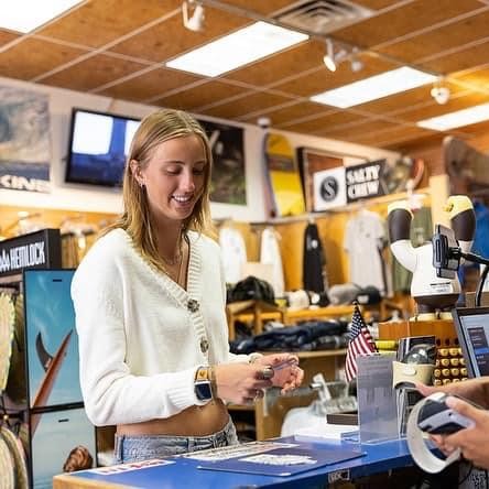 Una mujer de pelo rubio y jersey blanco sonríe mientras compra un artículo en un pequeño comercio. El dueño de un pequeño negocio con una gorra de béisbol negra le llama.