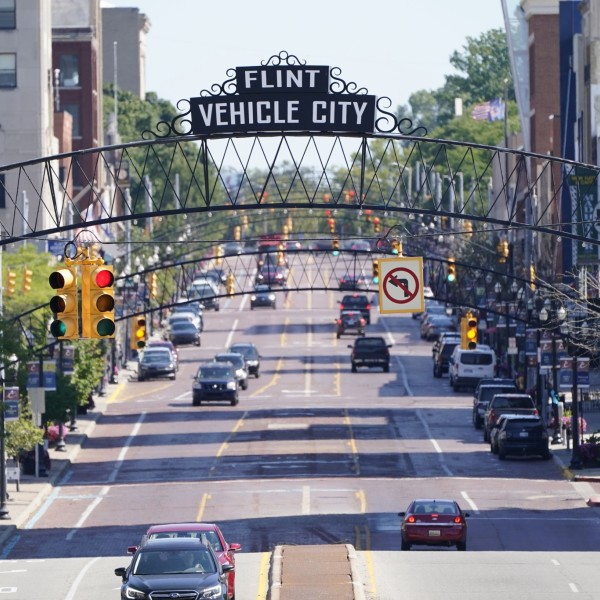 A street view of downtown Flint, Michigan, cars driving down the street, a sign reading, "Flint Vehicle City" is over the road.
