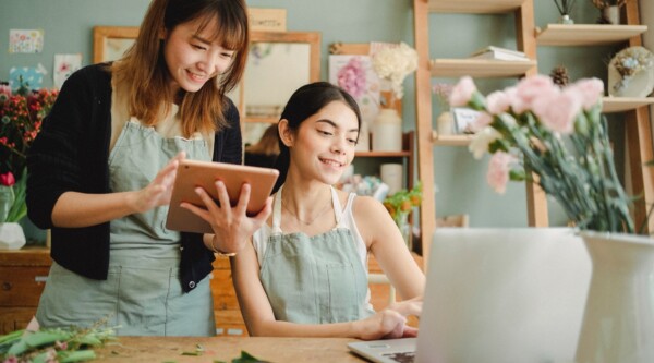 In a flower shop, two smiling women in aprons stand in front of a computer.