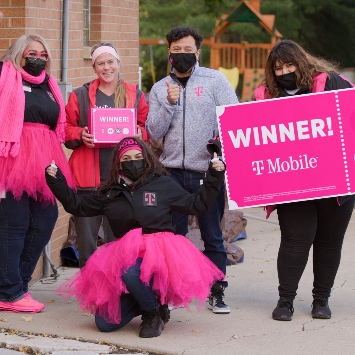 Un grupo de personas con accesorios de color rosa brillante, como tutús y bufandas, saludan y sonríen mientras sostienen un gran cheque rosa brillante que dice "¡Ganador!" con la marca T-Mobile.