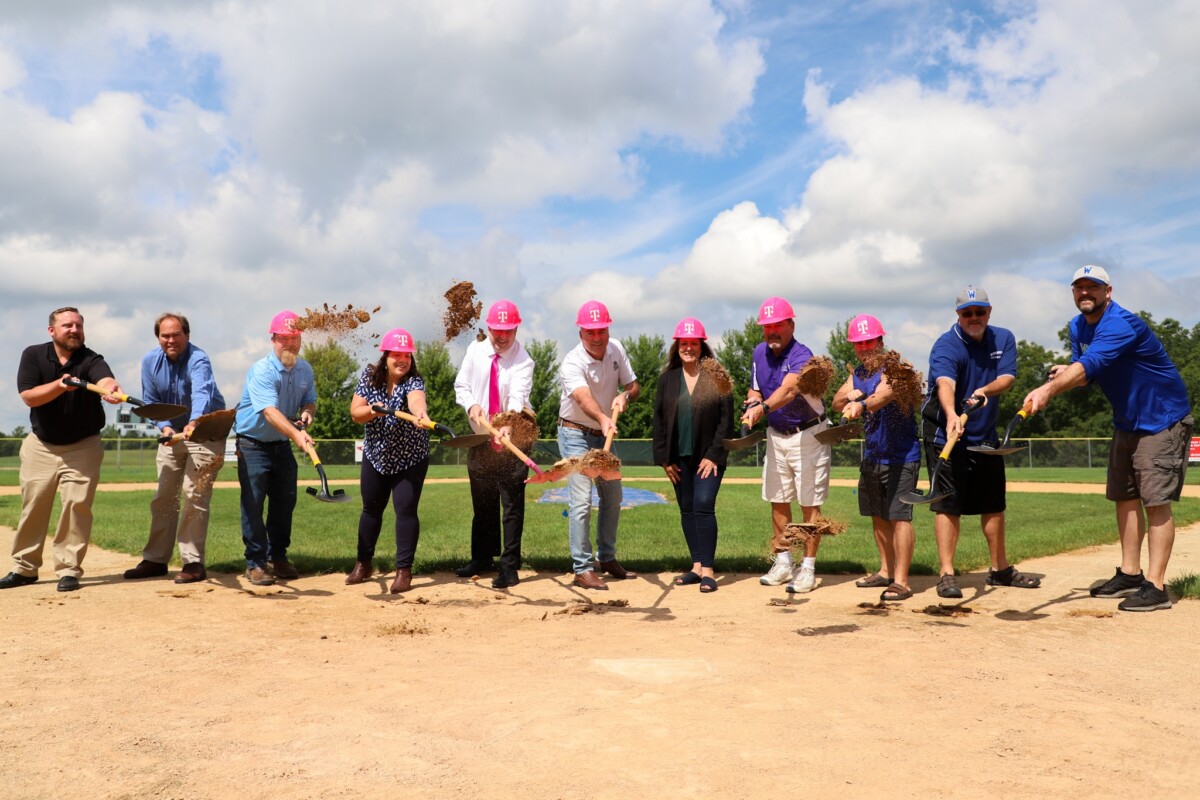 A group of people wearing T-Mobile branded hard hats and holding shovels gather on a baseball field.