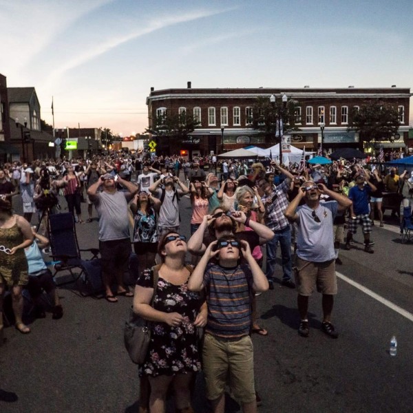 Photo of a crowd of people wearing eclipse protective eyeglasses look up at the sky in a downtown scene.