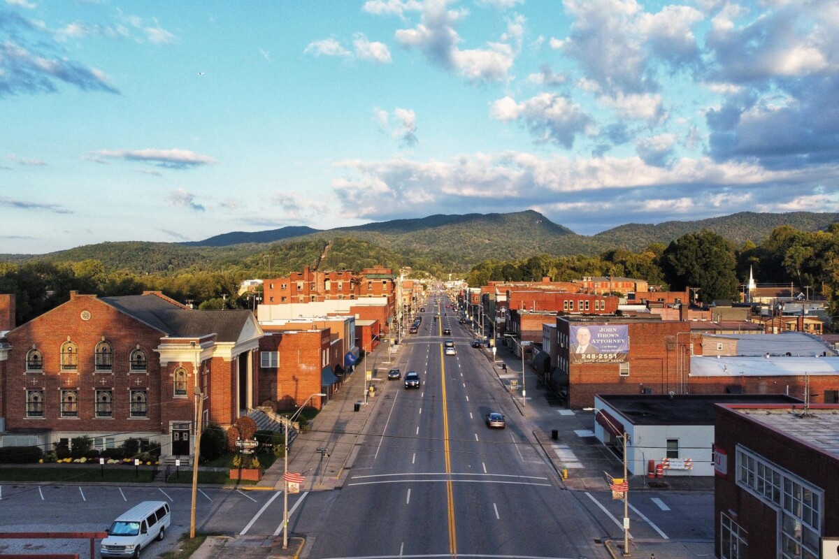 A photo of a downtown with brick buildings, four lane street, a hilly landscape, and a blue sky with clouds.
