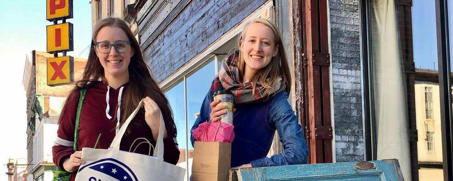 Two women hold local shopping bags outside of a small business. One tote bag reads, "Shop Small"