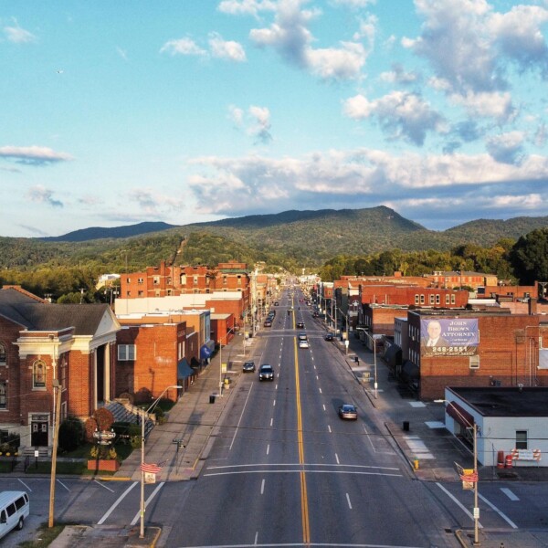 A photo of a downtown with brick buildings, four lane street, a hilly landscape, and a blue sky with clouds.