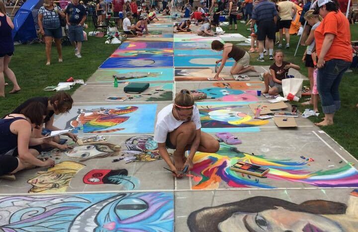 Spectators watch artists create chalk art on a sidewalk path.