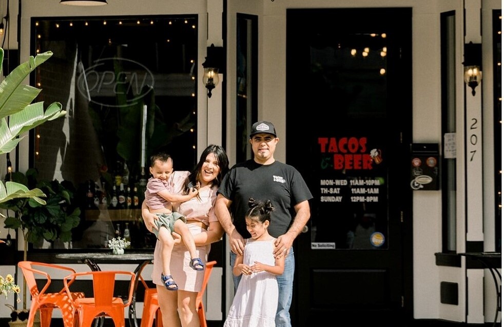 Una familia de dos adultos y dos niños pequeños posa delante de su negocio, Tacos & Beer.