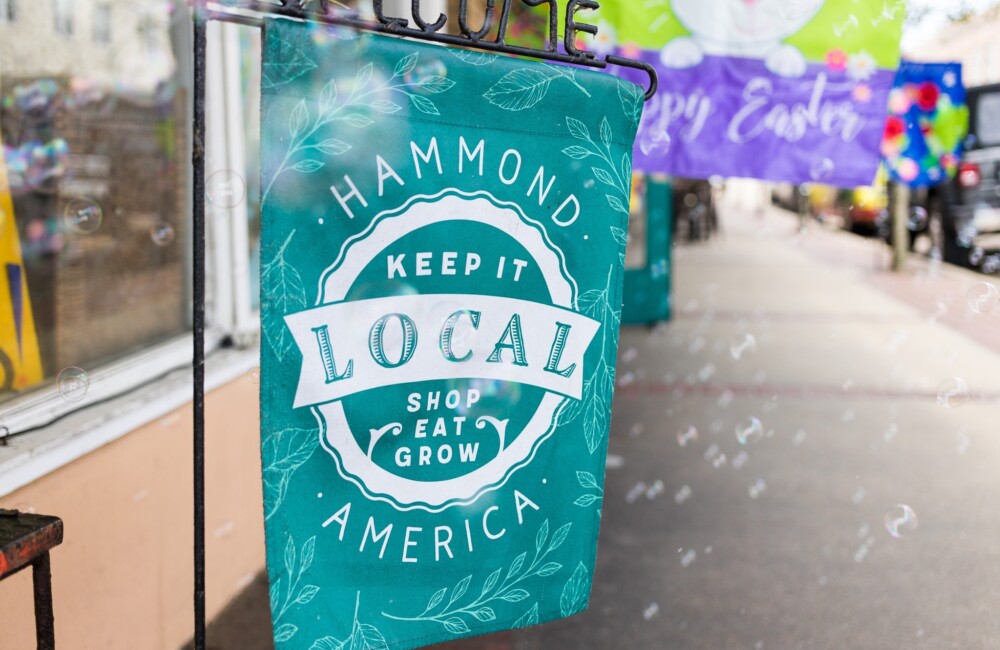 Una vibrante pancarta verde azulado cuelga de un soporte metálico negro colocado en una acera. La pancarta reza: "Hammond America: Keep it Local. Compra, come, cultiva".