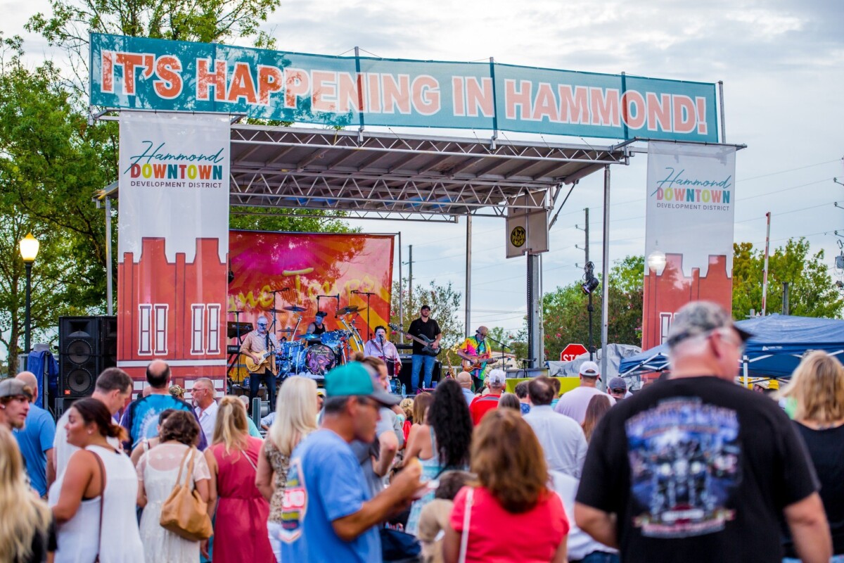 A large audience gathers in front of an outdoor stage where a band plays; a banner reading "It's Happening in Hammond" is stretched across the top of the stage.