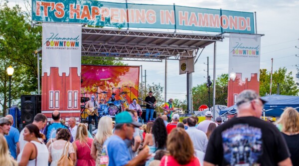 A large audience gathers in front of an outdoor stage where a band plays; a banner reading "It's Happening in Hammond" is stretched across the top of the stage.