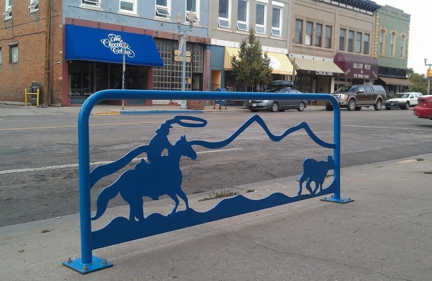 Aparcabicicletas de metal azul con una escena que representa a un vaquero a caballo persiguiendo a un ternero.