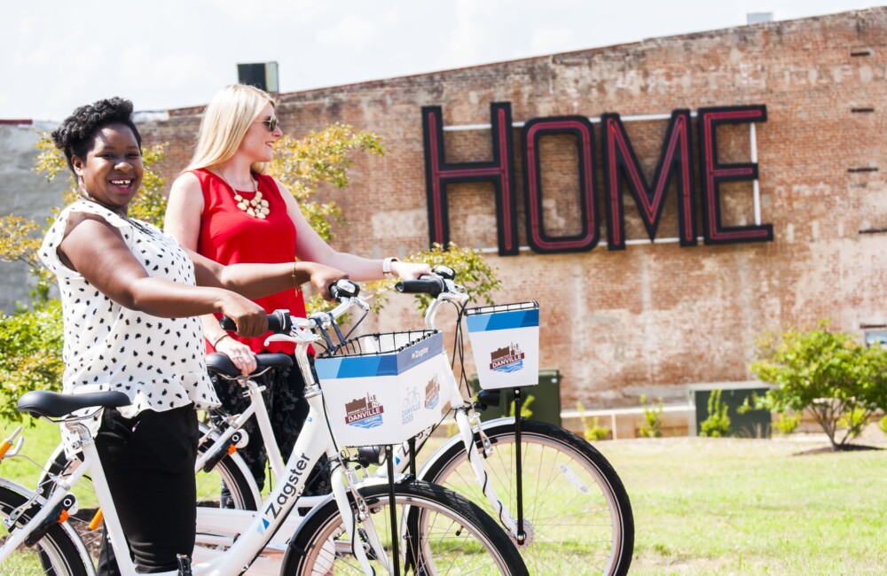 Dos mujeres sonríen mientras pasean bicicletas de alquiler por un parque de bolsillo bordeado por un edificio de ladrillo con la palabra "HOME" instalada en su exterior.