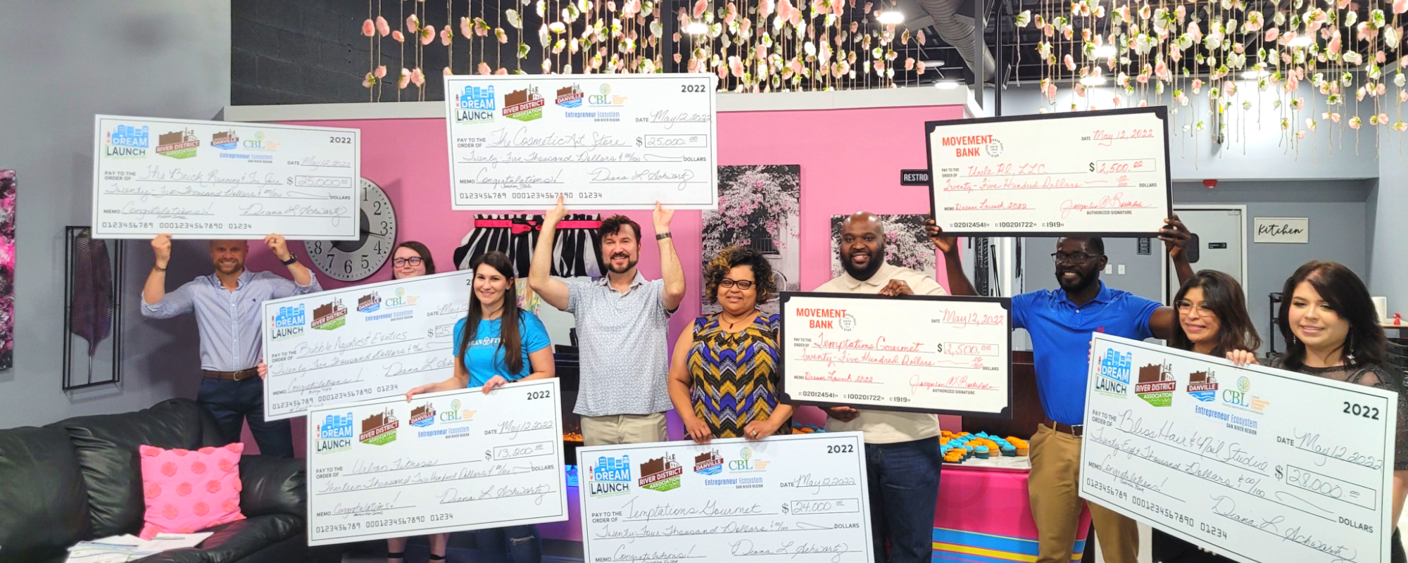 Un grupo diverso de nueve hombres y mujeres sostienen con orgullo cheques de gran tamaño y sonríen alegremente celebrando la concesión de capital inicial para apoyar sus iniciativas empresariales.