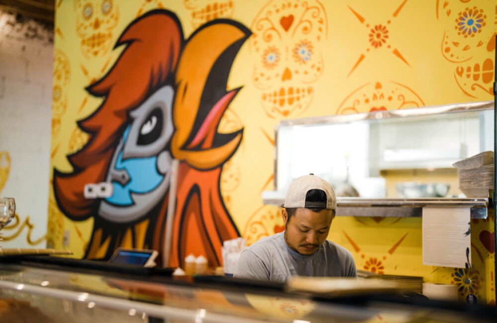 Un hombre trabaja detrás del mostrador de una charcutería, preparando un plato. Detrás de él se ve un vibrante mural con un gallo cacareando y una calavera mexicana.