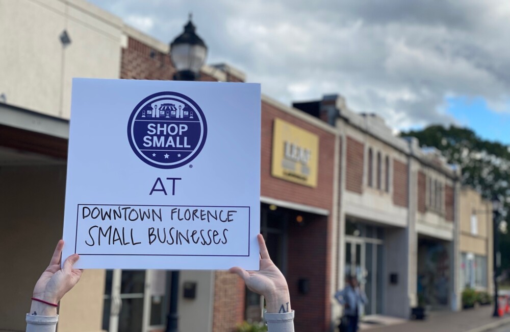 Un par de manos sostienen un cartel con el gráfico "Shop Small" y las palabras "At Downtown Florence Small Businesses" debajo. Al fondo se ven cinco escaparates con fachadas en buen estado.