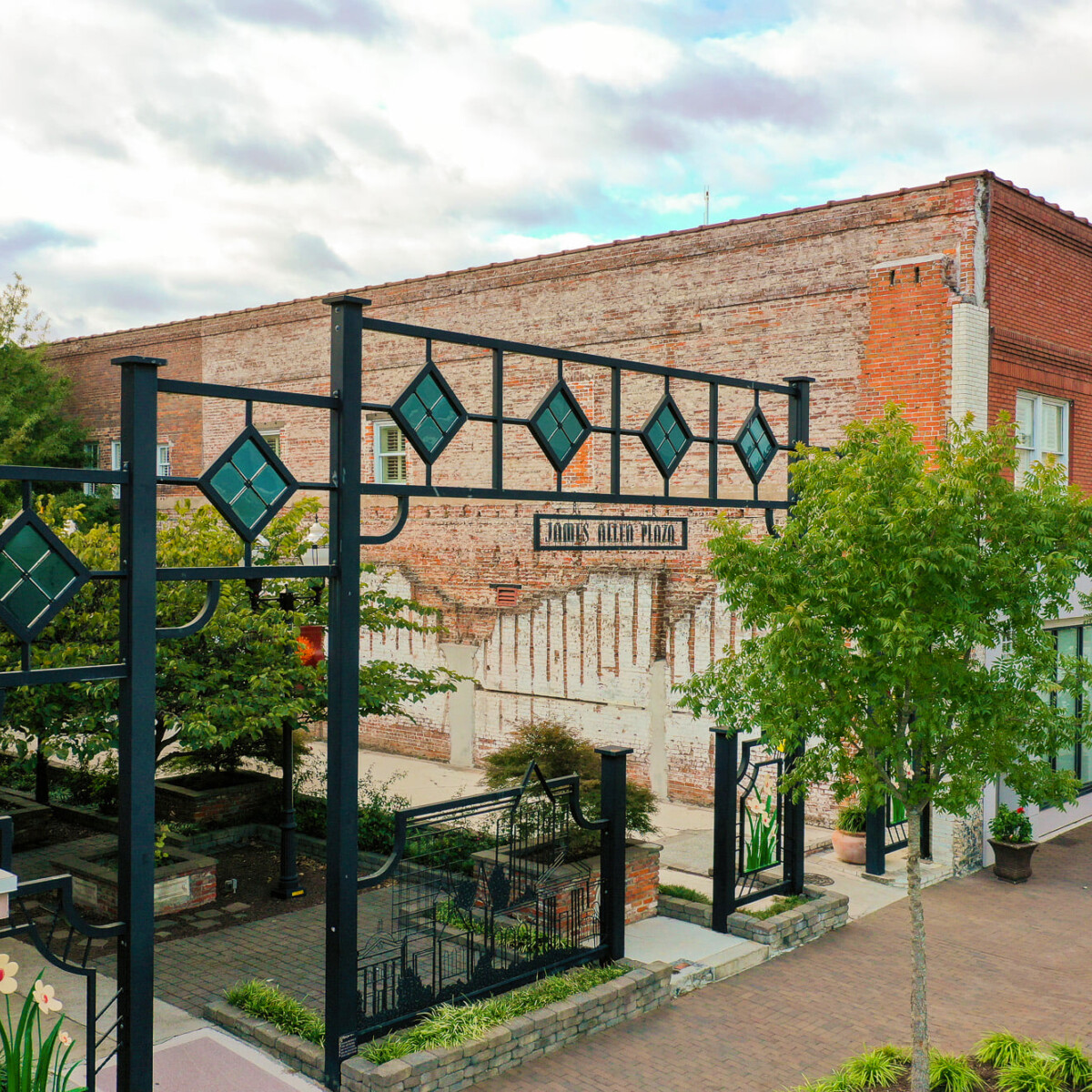 Two historic brick buildings flank the entrance to an outdoor plaza with a square, metal and stained glass gateway.
