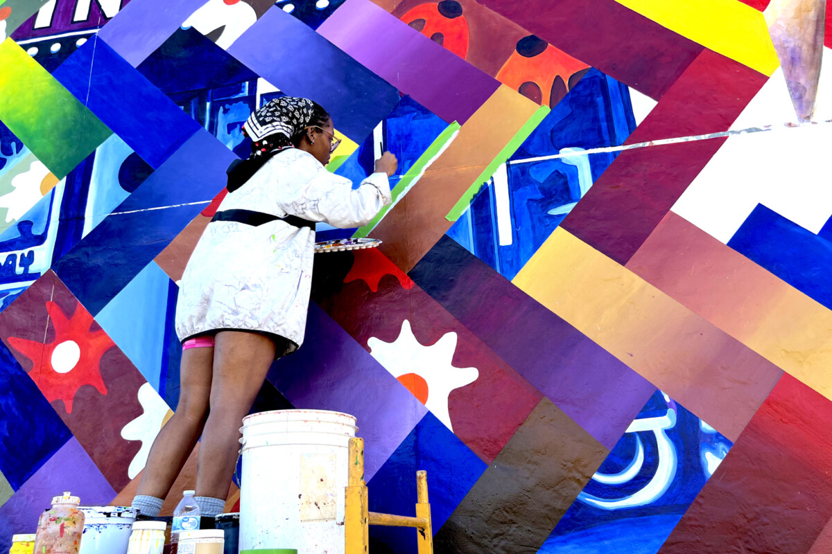 Un artista, de pie sobre un andamio, utiliza un pincel para aplicar pintura azul vibrante a una parte de un mural formado por un mosaico de líneas angulares entrelazadas, campos de color degradado y elementos decorativos.