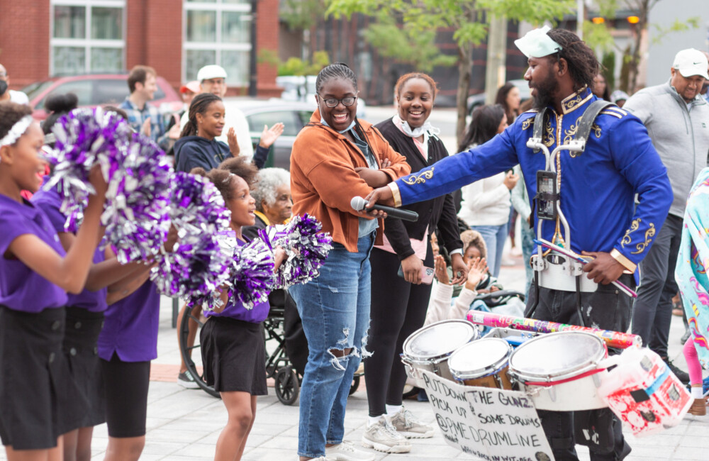 In a plaza, young cheerleaders dressed in purple and black shake pom poms while engaging with a snare drum musician who holds out a microphone to one of the cheerleaders.