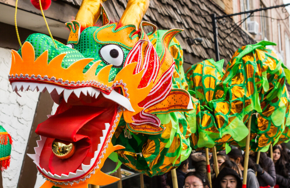 A large and brightly colored Chinese dragon during a Lunar New Year parade. Underneath the dragon, teenagers dressed in black with red sashes tied around their waists, walk in a single file line holding up poles attached to segments of the dragon.