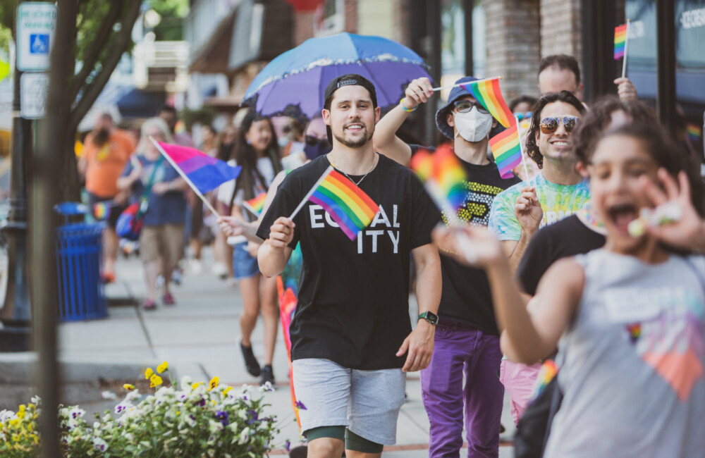 People participating in a Pride parade joyfully walk down a sidewalk while waving rainbow flags.