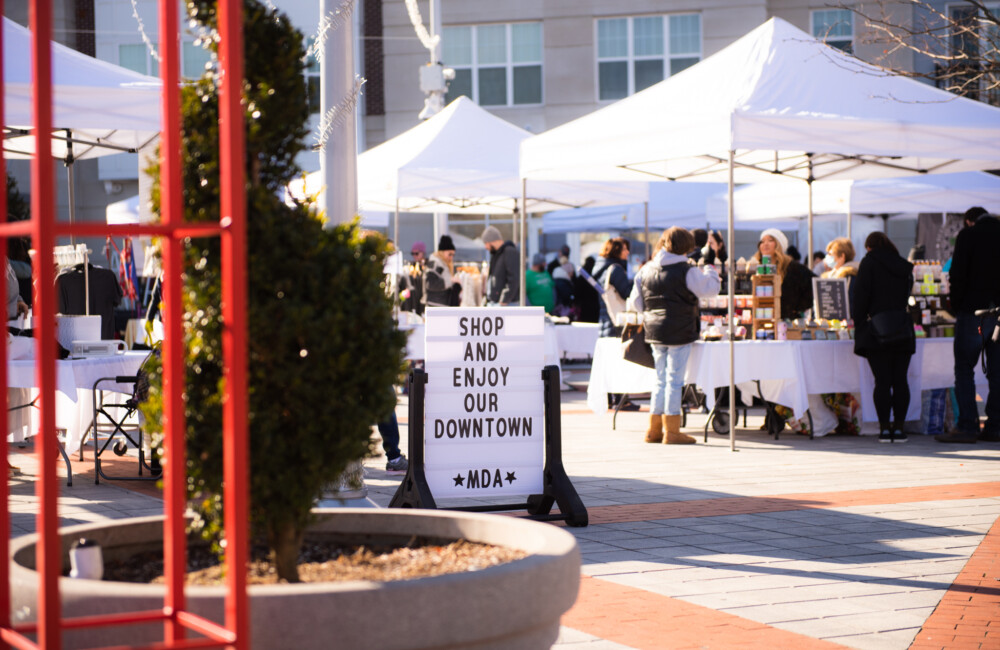 In a plaza, white pop-up canopy tents provide temporary storefronts for vendors at a winter market. In the foreground, a sign reads "Shop and Enjoy our Downtown - MDA" and in the background, the exterior of an new construction apartment complex is visible.