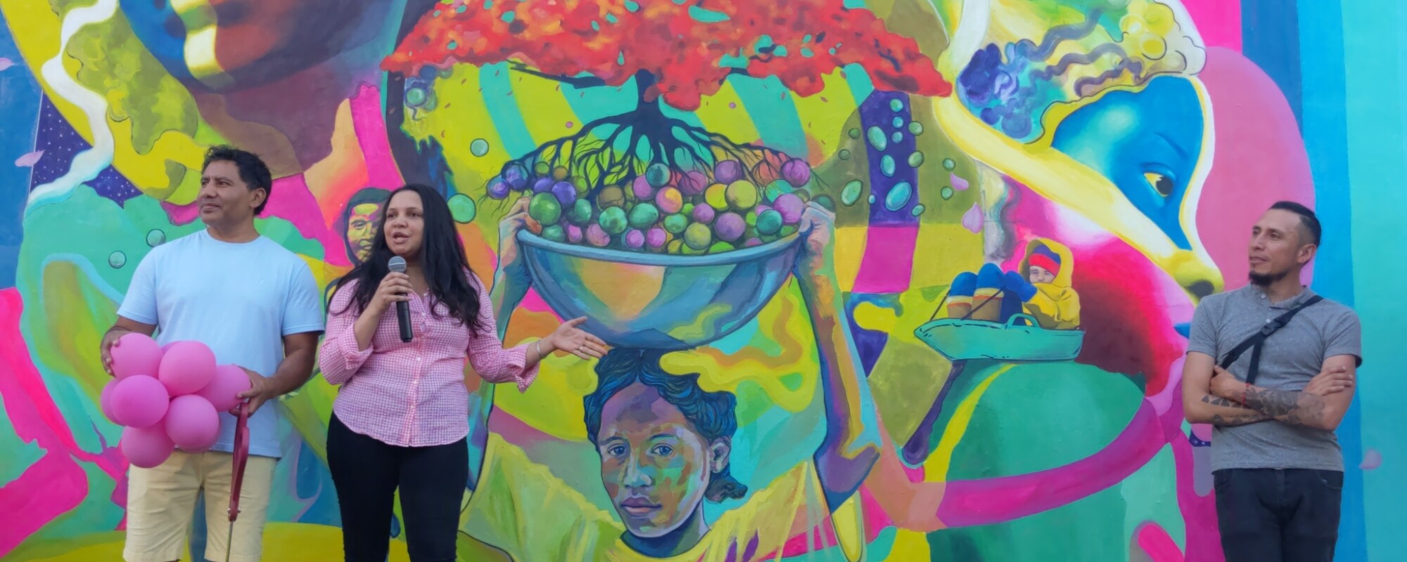 Two men and one woman stand in front of a brightly colored mural. The woman speaks into a microphone; to her right a man holds a cluster of balloons and a large pair of ceremonial scissors; the other man stands to her left.