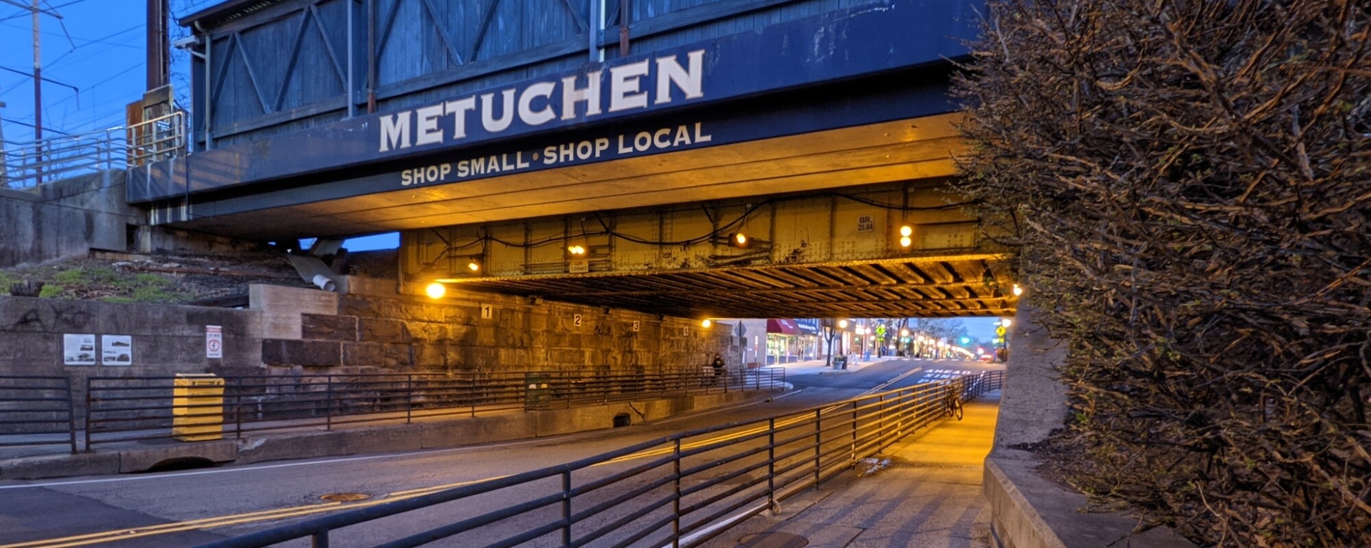 A covered, elevated railroad line crosses a roadway and pedestrian walkway. The words "Metuchen Shop Small, Shop Local" are painted in large white letters.