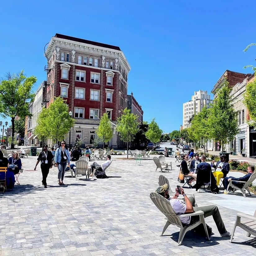 People lounge in chairs, sit at bistro tables, and stroll through a large plaza dotted with trees and surrounded by historic brick buildings.