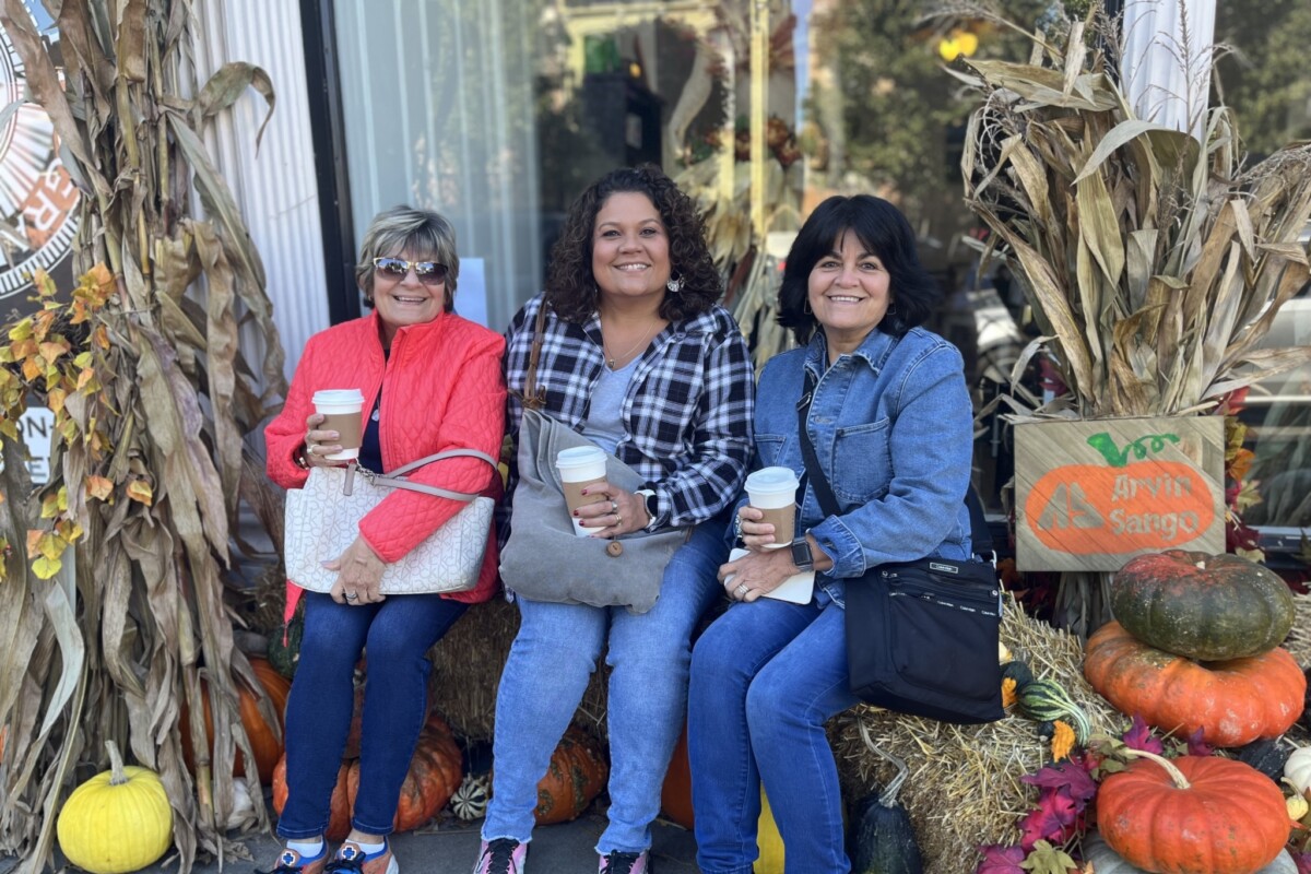 Three women hold cups of coffee while sitting on a hay bale.