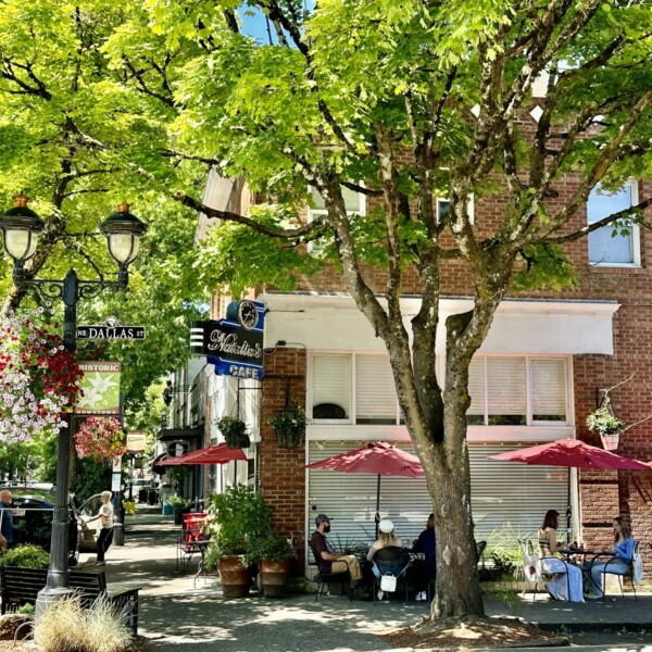 En un día soleado, los comensales disfrutan de los asientos al aire libre bajo la sombra de los árboles y de las comidas de una cafetería situada en un histórico edificio de ladrillo.