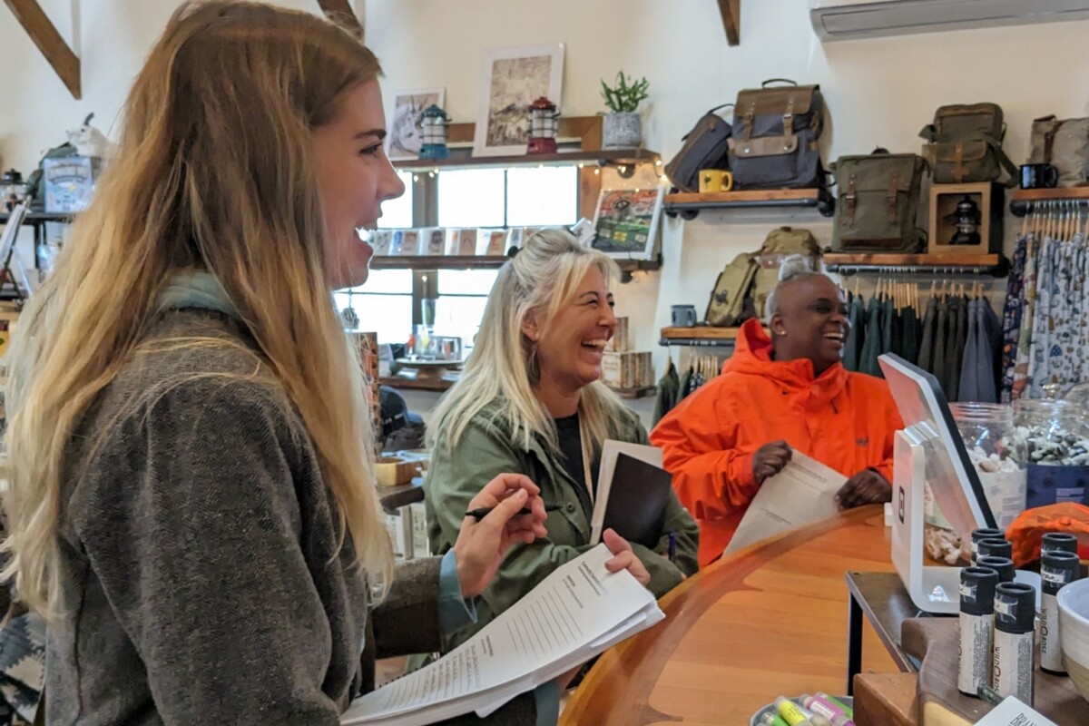 Three women stand inside a gift shop and laugh while speaking with the shop clerk.