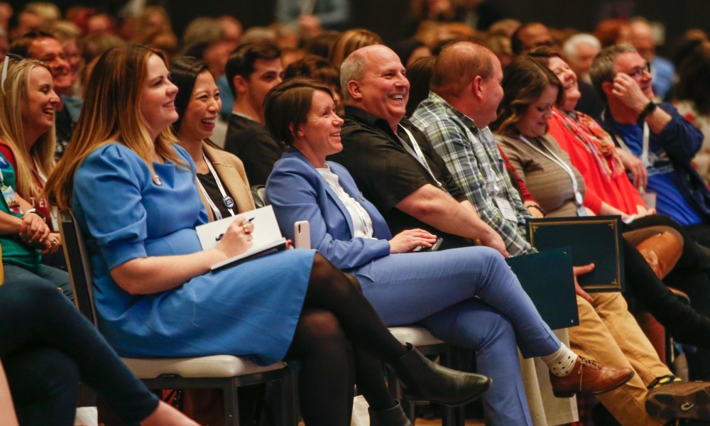 Gente sentada al estilo de un teatro en una gran sala, riendo junto a un orador (que no aparece en la foto).