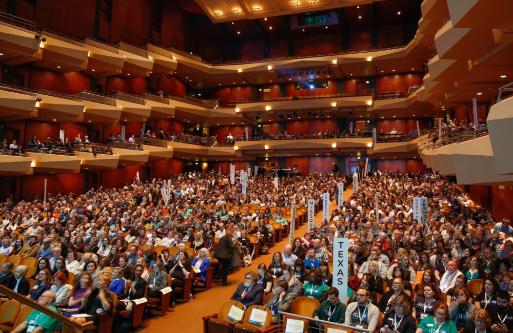 Over 1000 people gather in a large auditorium with theater and balcony seating, some hold signs displaying the names of U.S. states.
