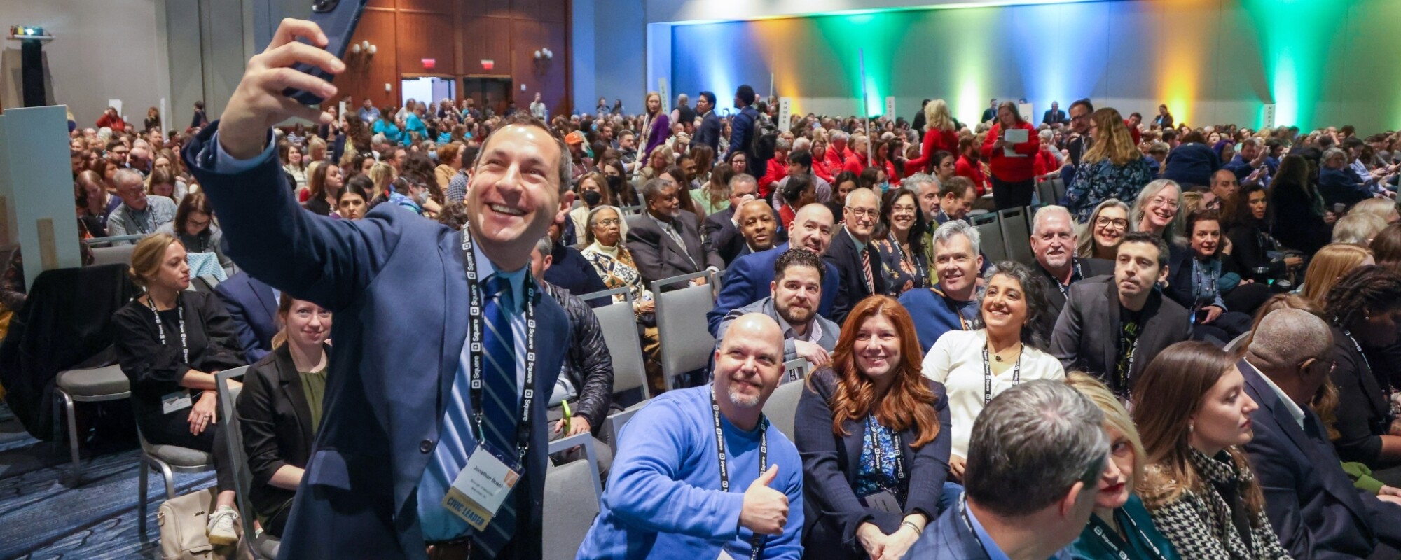 In a large conference ballroom, people find seats while a man takes a selfie with colleagues.