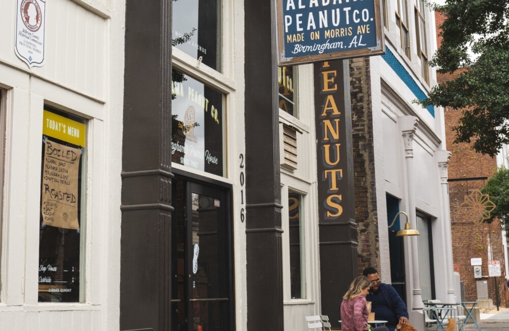 Dos personas se sientan a una mesa en la acera frente a una tienda situada en un edificio histórico de ladrillo. El cartel que cuelga del exterior del edificio reza "Alabama Peanut Co.".