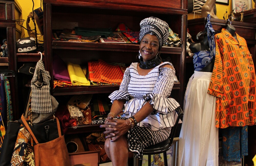 Una mujer negra se sienta en una silla y sonríe rodeada de la colorida ropa africana expuesta en su tienda.