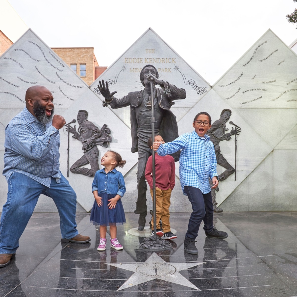 Un padre y sus tres hijos pequeños cantan y bailan mientras visitan el monumento a Eddie Kendrick; al fondo se ven estatuas de bronce del cantante.
