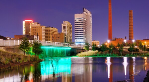 La parte inferior de un puente se ilumina con luces de color verde azulado; el perfil de la ciudad y las chimeneas de color óxido que se elevan en el fondo se reflejan en el río que fluye suavemente en primer plano.