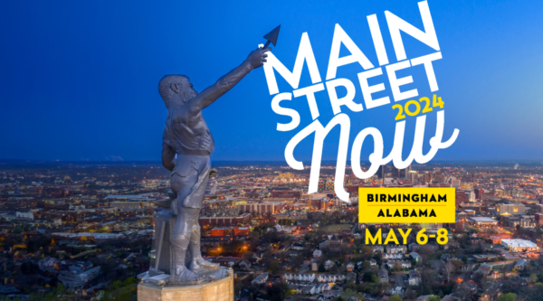 Estatua de Vulcano con el horizonte iluminado de la ciudad de Birmingham al fondo. La lanza alzada de la estatua señala el logotipo de Main Street Now 2024 en la esquina superior derecha.