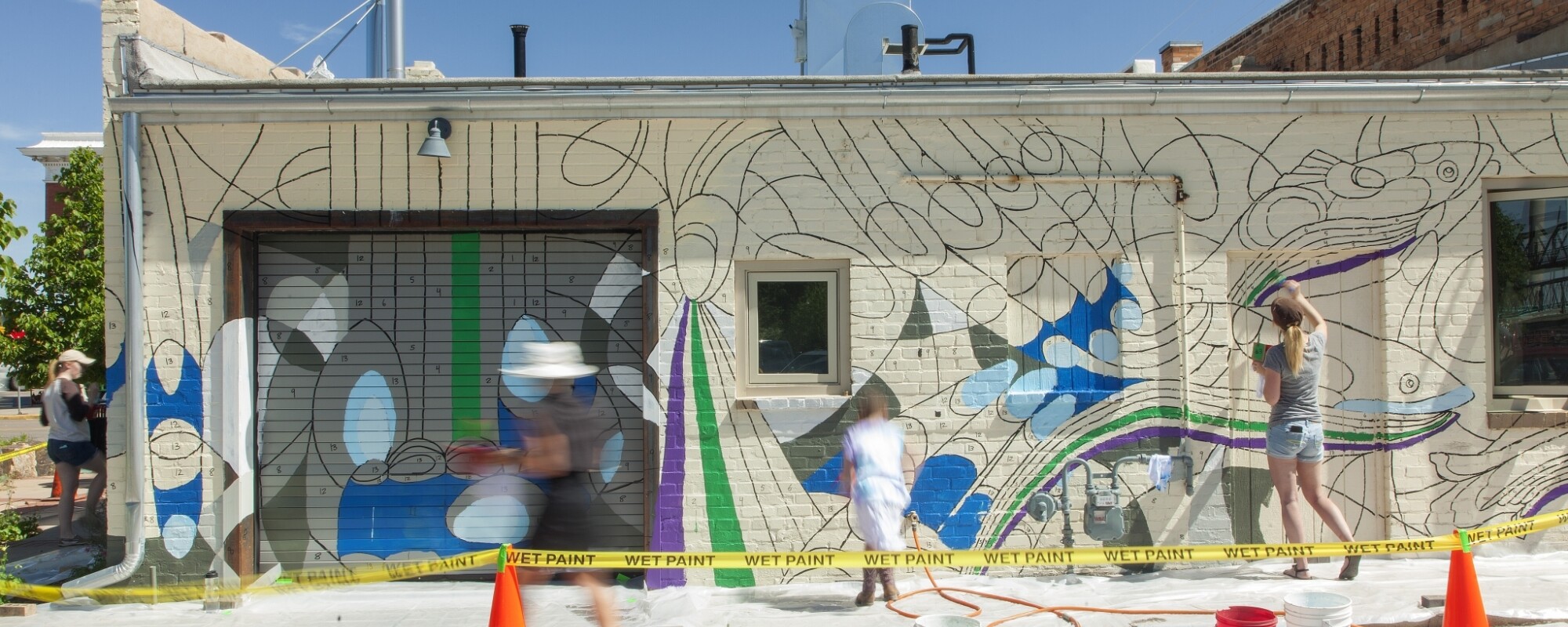 People helping paint a mural with different shades of blue on the side of a downtown building.