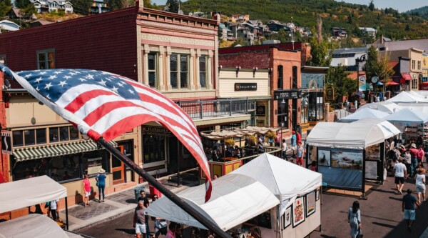 La histórica Main Street durante una feria callejera con vendedores y gente paseando; una gran bandera estadounidense ondea en primer plano.