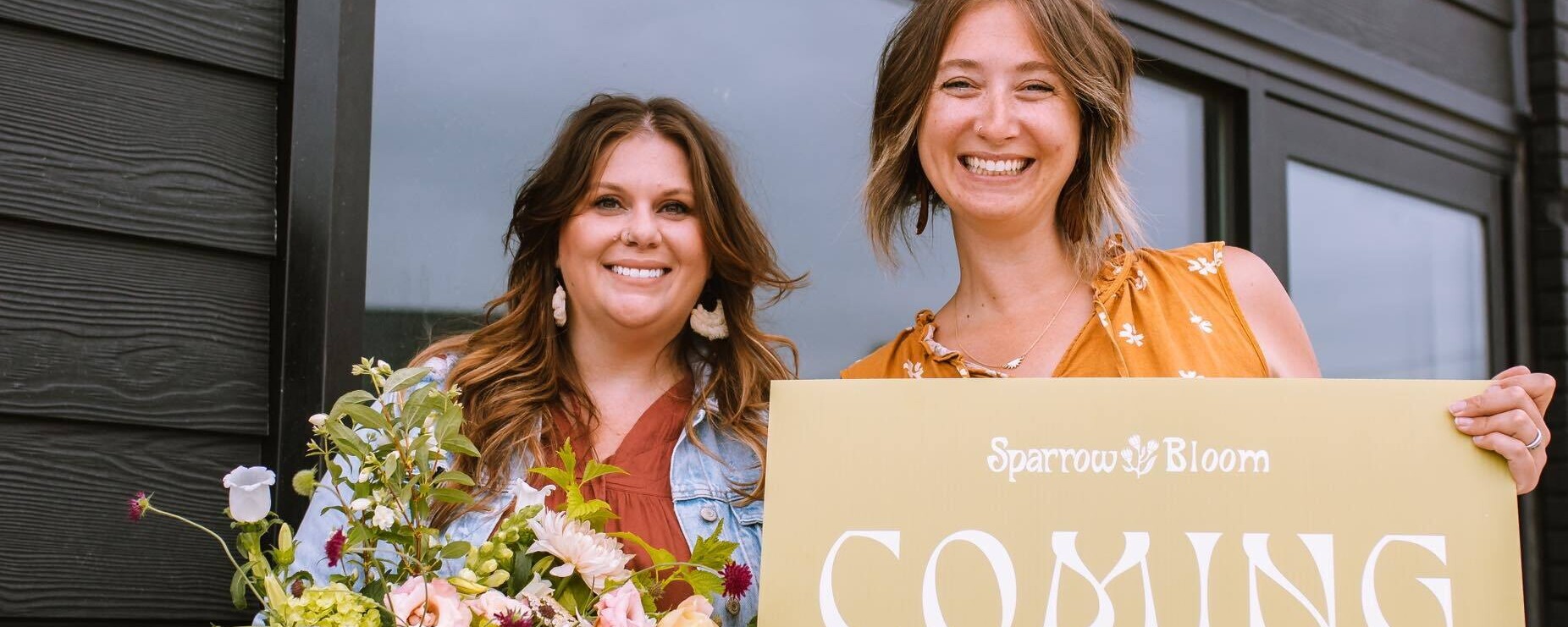 Dos mujeres posan delante de su nuevo escaparate: una sostiene un ramo de flores mientras la otra sostiene un cartel en el que se lee "¡Próxima apertura!".