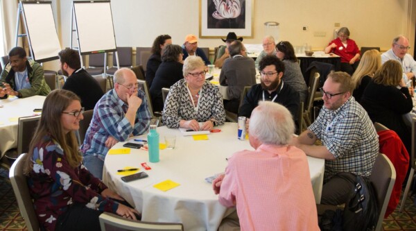 Seis personas se sientan alrededor de una mesa para discutir los materiales del curso. Al fondo, otros participantes discuten en pequeños grupos.