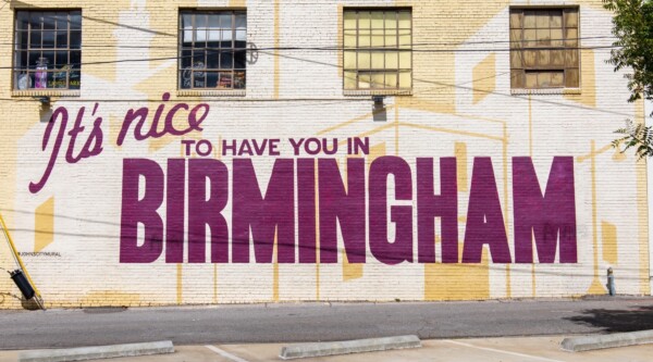 Mural on the exterior wall of a building reads "It's Nice to Have you in Birmingham" in a deep magenta color.
