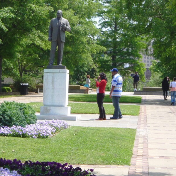 La gente pasea por un parque bellamente ajardinado mientras un hombre y una mujer se detienen a visitar una estatua de bronce de Martin Luther King, Jr.