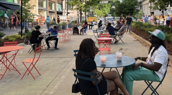 People sitting at café tables set-up along a street lined with tall office buildings.