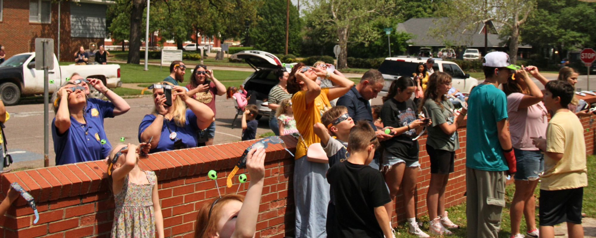 People standing by a brick wall along a street look at the sun through eclipse glasses