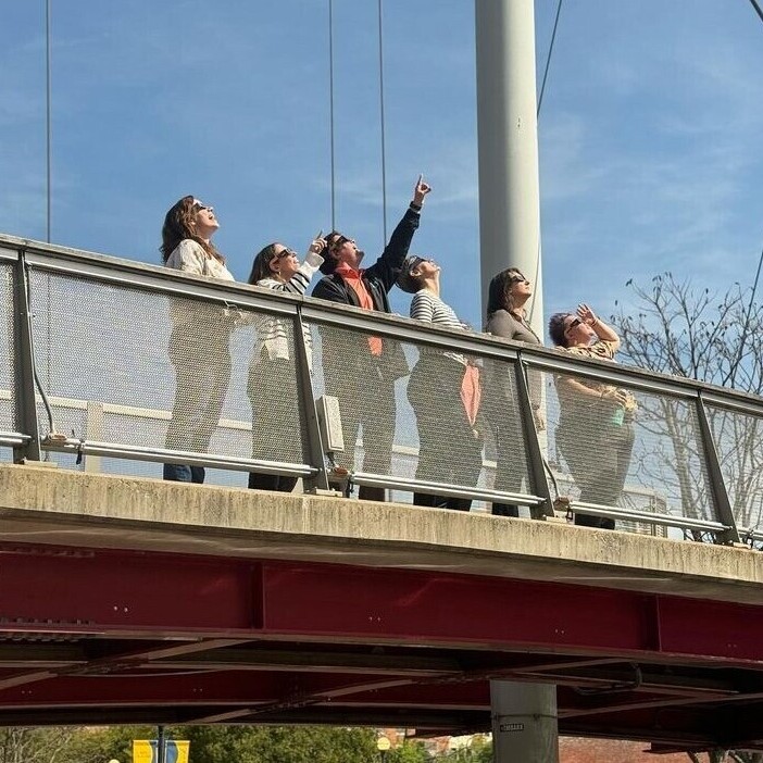 People standing on a bridge look up at the sky, one man points at the sun