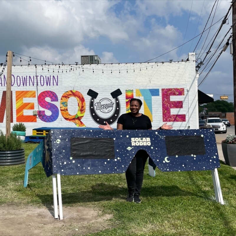 Tia Vice poses behind a pair of giant eclipse glasses in front of a mural reading "#IAmDowntownMesquite