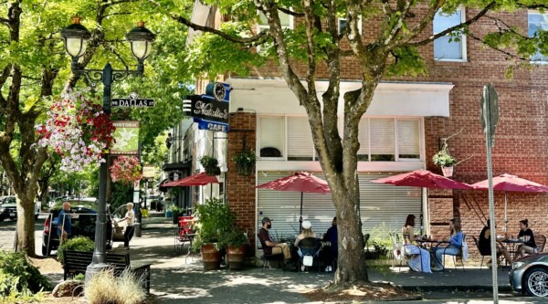 On a sunny day, diners enjoy outdoor sidewalk seating under the shade of trees and meals from a café located in a historic brick building.